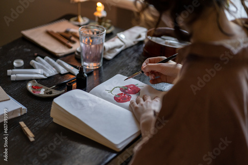 Creative hobbies. Close up of female artist sitting at table with burning candles painting in sketchbook at home or studio, using oil paints. Young woman talented painter creating new work of art