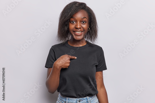 Fotografi Surprised cheerful Afro woman points at herself asks who me dressed in casual black t shirt smiles pleasantly isolated over white background