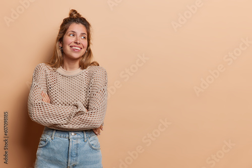 Stunning and cheerful woman keeps arms folded looks aside recalls something funny dressed in casual knitted jumper jeans isolated over brown background copy space for youfr advertising content © wayhome.studio 