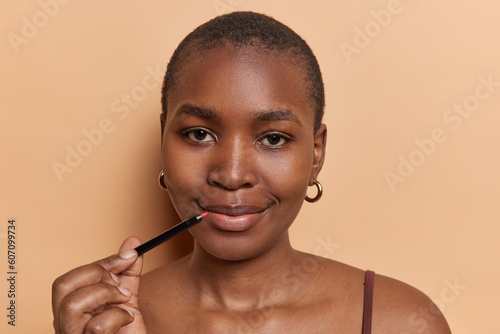 Horizontal shot of short haired black woman holds red lip pencil puts on makeup dressed in casual t shirt looks directly at camera isolated over brown background. Afro female draws lips with lipliner