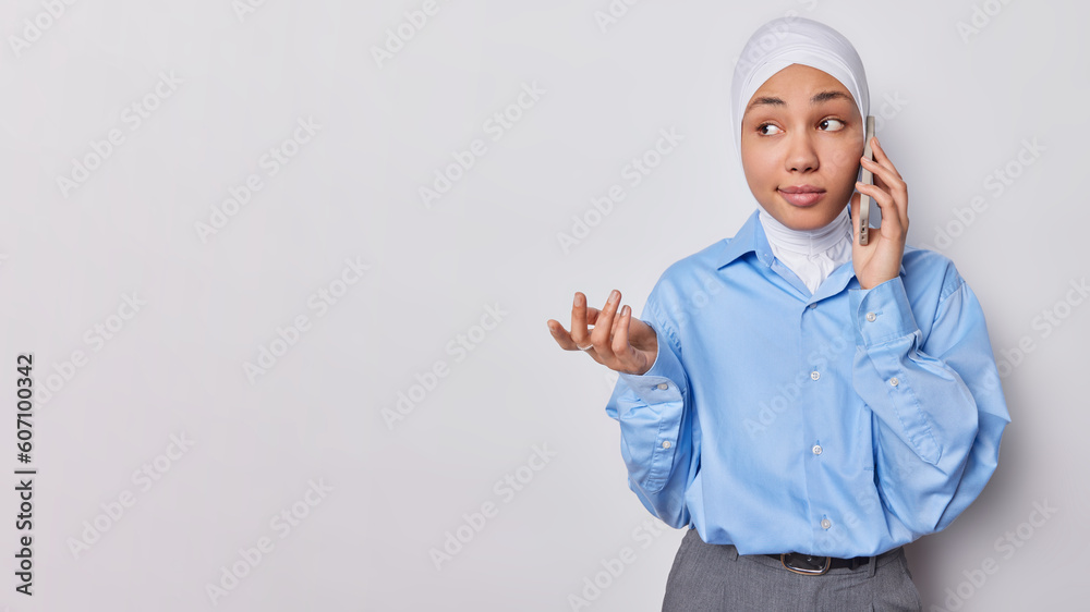 Horizontal shot of confused Muslim woman has phone conversation shrugs shoulders feels uncertain wears traditional veil and formal outfit isolated on white background copy space for your advertisement Horizontal shot of confused Muslim woman has phone conversation shrugs shoulders feels uncertain wears traditional veil and formal outfit isolated on white background copy space for your advertisement