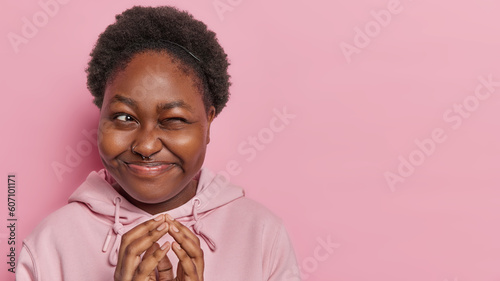 Mysterious dark skinned woman has mischievous grin steeps fingers looks with cunning expression being intrigues dressed in casual sweatshirt poses against pink background. Hmm let me think about it