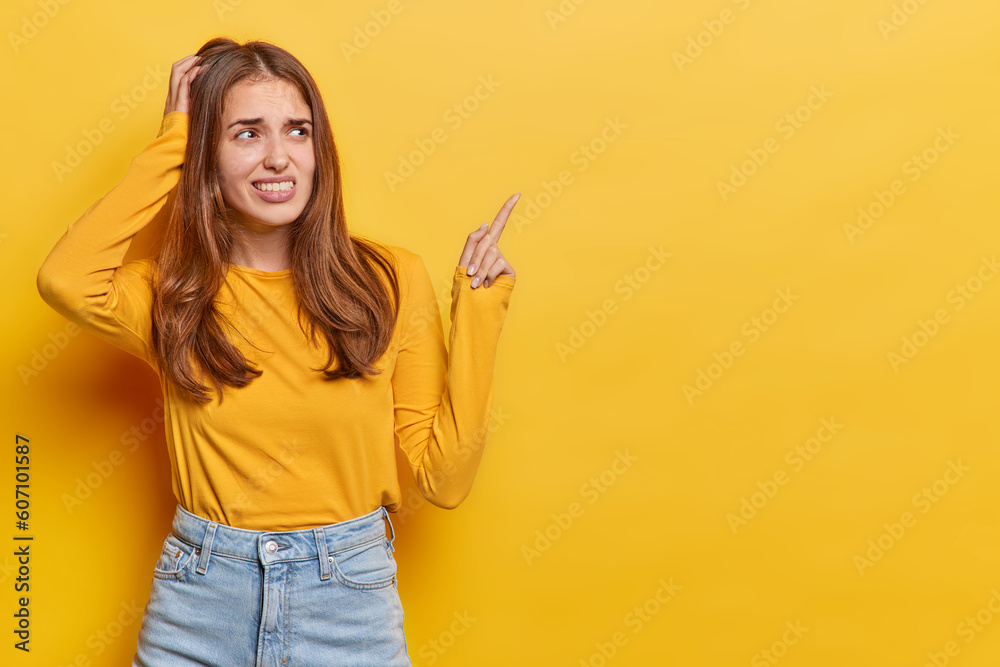 Horizontal shot of displeased puzzled woman scratches head clenches teeth points index finger at copy space dressed in casual jumper and jeans isolated over yellow background. Advertisement concept Horizontal shot of displeased puzzled woman scratches head clenches teeth points index finger at copy space dressed in casual jumper and jeans isolated over yellow background. Advertisement concept