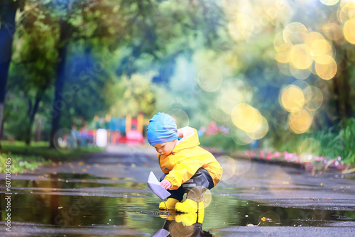 Fototapeta Naklejka Na Ścianę i Meble -  paper boat puddle game, boy seasonal autumn look raincoat, yellow rubber boots, outdoor game