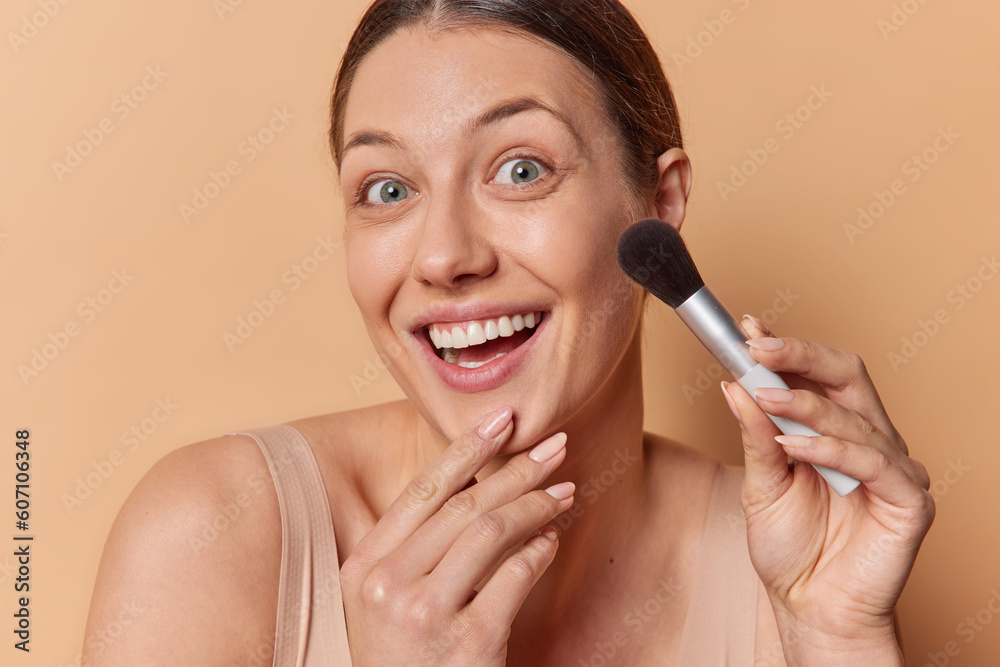 Headshot of smiling brunette adult woman applies blush on cheekbones with cosmetic brush keeps hand on chin smiles toothily poses indoor against brown background gest ready prepare in morning Headshot of smiling brunette adult woman applies blush on cheekbones with cosmetic brush keeps hand on chin smiles toothily poses indoor against brown background gest ready prepare in morning