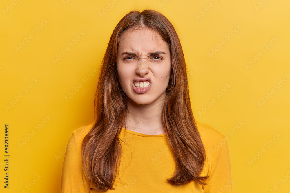 Portrait of dark haired young European woman keeps teeth clenched frowns face being irritated dressed in casual jumper isolated over vivid yellow background. People irritation and anger concept Portrait of dark haired young European woman keeps teeth clenched frowns face being irritated dressed in casual jumper isolated over vivid yellow background. People irritation and anger concept