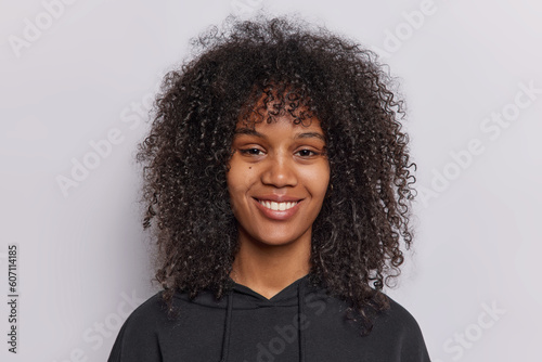 Portrait of happy curly haired teenage girl smiles pleasantly looks directly at camera dressed in casual black sweatshirt isolated over white background beams with joy has genuine smile on face