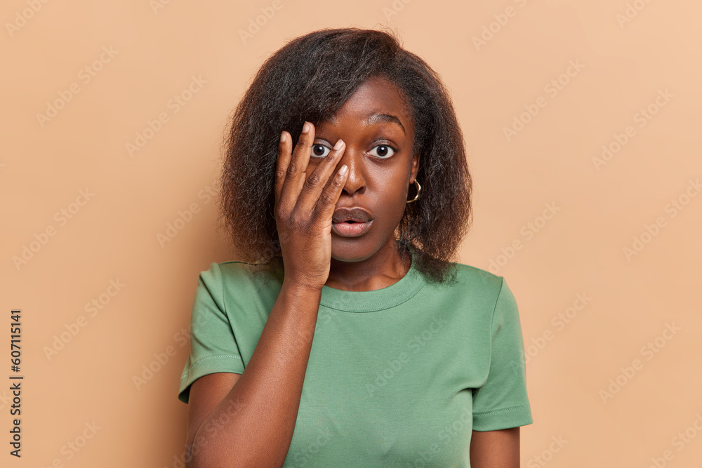 Shocked dark skinned woman covers half of face stares with scared expression reacts to something terrible dressed in casual green t shirt isolated over brown background. Reaction to something horrible Shocked dark skinned woman covers half of face stares with scared expression reacts to something terrible dressed in casual green t shirt isolated over brown background. Reaction to something horrible