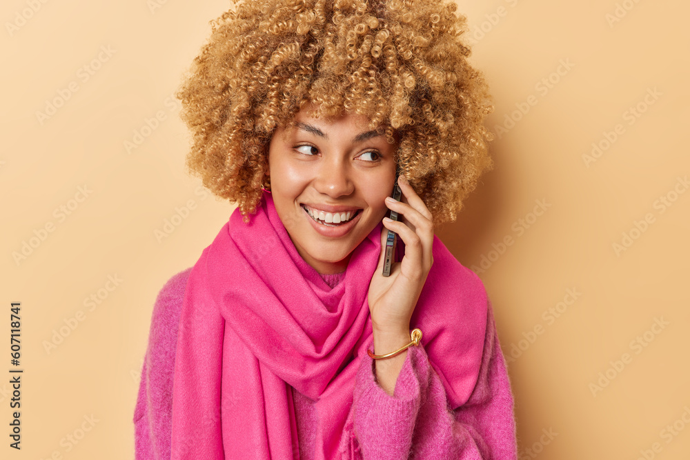 Lovely young woman holds phone near ear has meaningful conversation receives pleasant news smiles happily concentrated aside wears pink jumper and scarf isolated over brown studio background Lovely young woman holds phone near ear has meaningful conversation receives pleasant news smiles happily concentrated aside wears pink jumper and scarf isolated over brown studio background