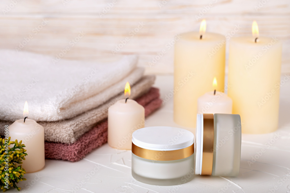 Jars with day and night face cream, burning candles, towels and juniper branch on light background. Concept of calmness, comfort, spa treatments. Selective focus