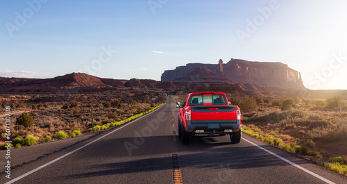 Red Pickup Truck driving on Scenic Road in the Dry Desert with Red Rocky Mountains in Background