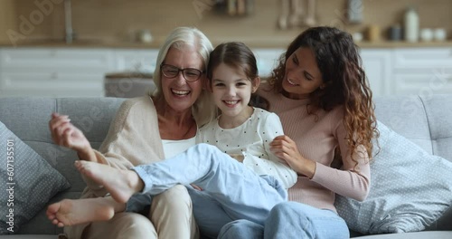 Cheerful grandmother and young mom looking at camera play with little kid on cozy couch in living room, tickle girl, have fun together at home, enjoy carefree weekend. Family ties, playtime activity photo