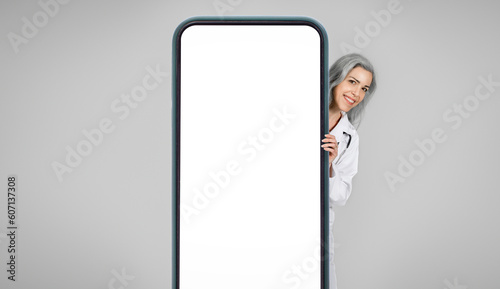 Happy Doctor Woman Peeking Behind Large Smartphone Screen, Gray Background © Prostock-studio