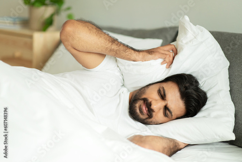 Despaired sad millennial middle eastern guy covers ears with pillow, suffers from noise and insomnia, lies on bed