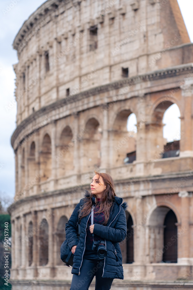 Pensive young Latin woman tourist in warm clothes and professional camera standing against Colosseum and looking away while admiring historic architecture during trip through Rome, Italy