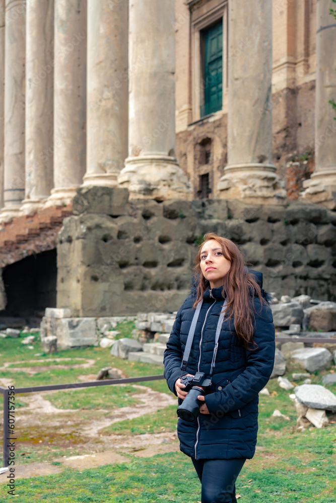 Pensive young Latin woman tourist in warm clothes and professional camera standing in Roman Forum next to Colosseum and looking away while admiring historic architecture during trip in Rome, Italy
