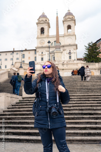 Trendy young Latin woman tourist in sunglasses and warm clothes standing with smartphone and professional photo camera looking away in Piazza di Spagna in Rome, Italy