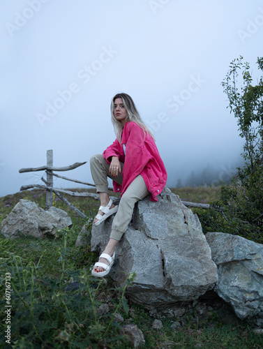 A girl in a pink jacket sitting on a stone. A mysterious environment with the fog behind it.