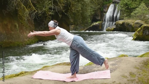 Mature woman is practicing yoga on a rocky beach by the river. Waterfall photo