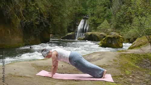 Mature Woman Practicing Yoga on a beach by the river in front of a waterfall photo