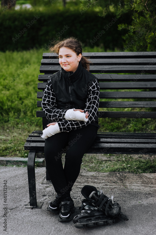 Fototapeta premium A girl in sadness, a child athlete with a cast, a bandage on her hands after a bone fracture, sits on a bench in the park with roller skates, missing riding alone. Photography, portrait, sports.