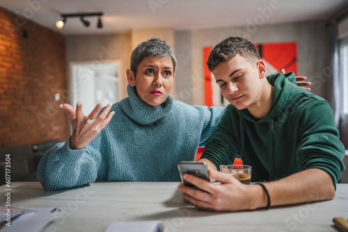 teenage boy and mature caucasian woman sit together at the kitchen at home talk mother and son or relatives support solving problem share experience and opinion boy hold mobile phone smartphone