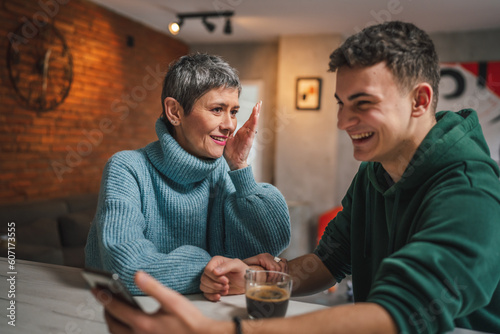 teenage boy and mature caucasian woman sit together at the kitchen at home talk mother and son or relatives support solving problem share experience and opinion boy hold mobile phone smartphone