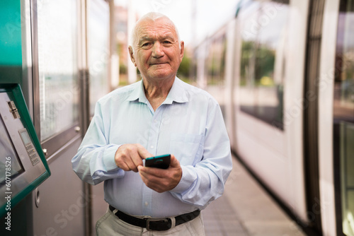 Old man standing on tram station and using smartphone while waiting.