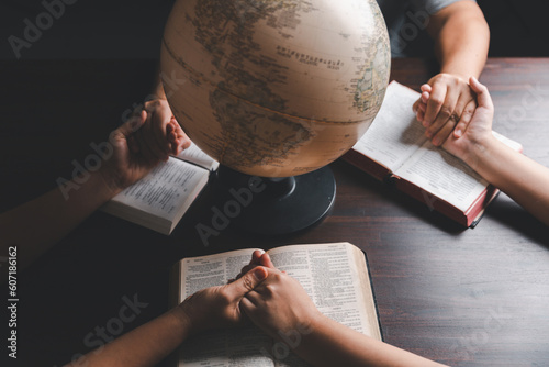 Christian group praying for globe and people around the world on wooden table with bible. Christian small group holding hands and praying together around a wooden table with bible page in homeroom.