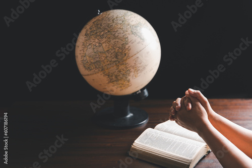 Christian woman praying for globe and people around the world on wooden table with bible. Christian hands praying together around a wooden table with bible page in homeroom.