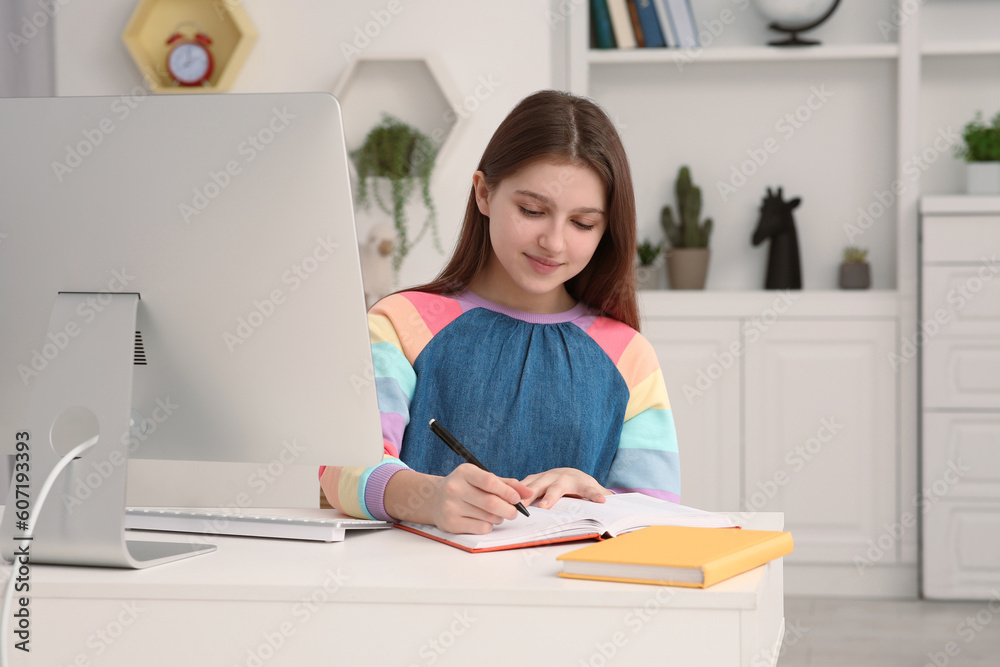 Cute girl writing in notepad while using computer at desk in room. Home workplace Cute girl writing in notepad while using computer at desk in room. Home workplace