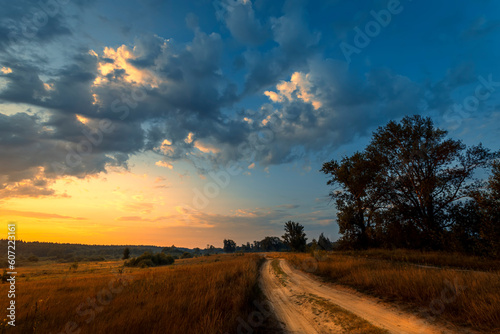 Rural road in green grass and orange summer sunrise or sunset.