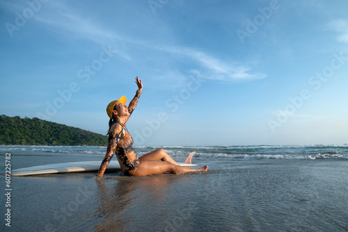 Surfer girl sits on the beach in front of the sea and clear blue sky, feeling happy waving and looking at the sky, outdoor activities, water sports activities concept