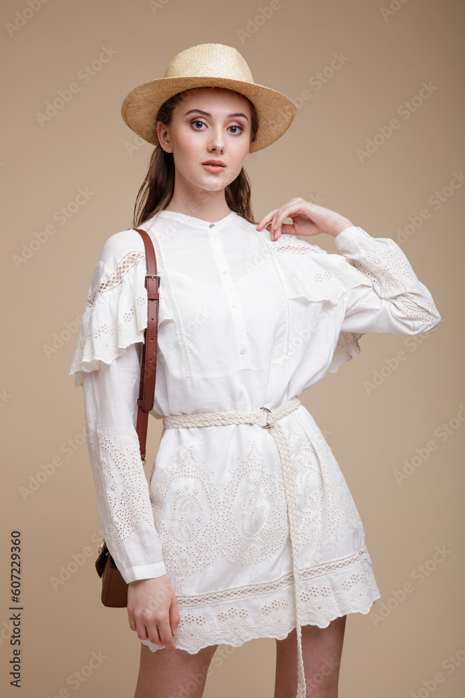 High fashion photo of beautiful elegant young woman in a pretty white dress with the sleeves, hat, handbag posing over beige brown background. Studio Shot, portrait. High fashion photo of beautiful elegant young woman in a pretty white dress with the sleeves, hat, handbag posing over beige brown background. Studio Shot, portrait.
