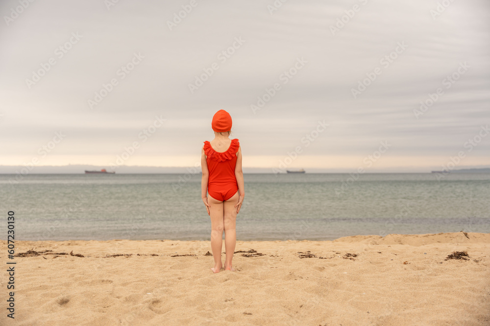 A little girl in a red bathing suit, in a red cap, walks along the sandy beach on the seashore, ocean. Looks at the sea. Sea holidays, travel and beach holidays with children