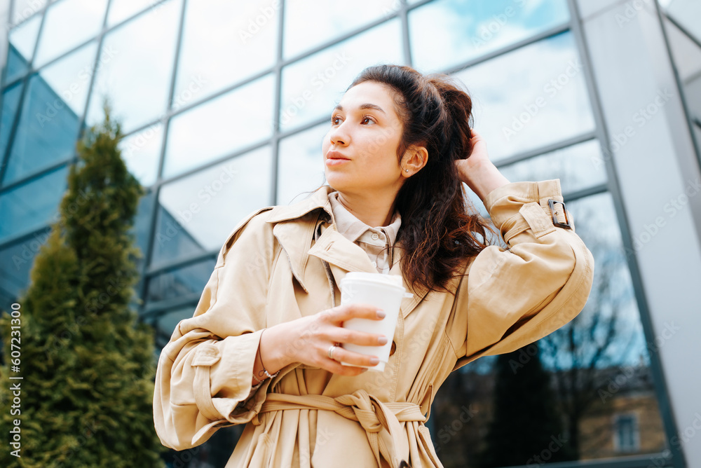 Stylish mixed race woman in raincoat holding disposable cup of coffee fixing hair while standing against modern glass building background, urban lifestyle