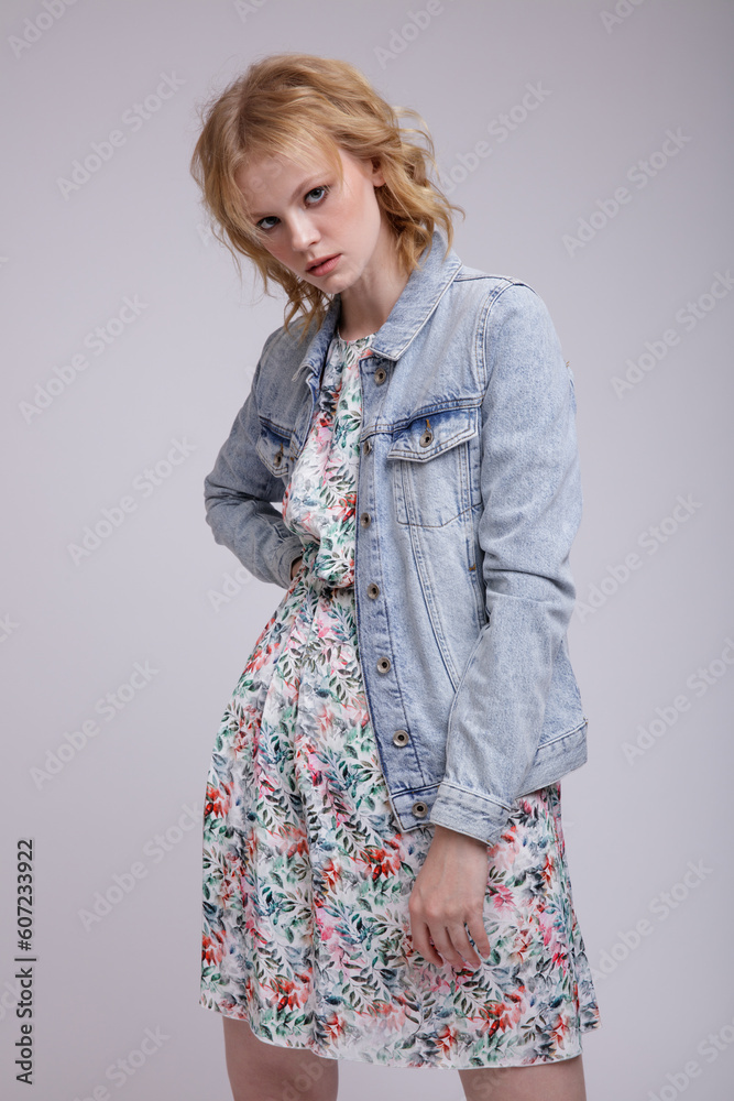 High fashion photo of beautiful elegant young woman in a pretty colorful dress with floral pattern, denim, jeans jacket posing over white, soft gray background. Studio Shot, portrait. Blonde High fashion photo of beautiful elegant young woman in a pretty colorful dress with floral pattern, denim, jeans jacket posing over white, soft gray background. Studio Shot, portrait. Blonde