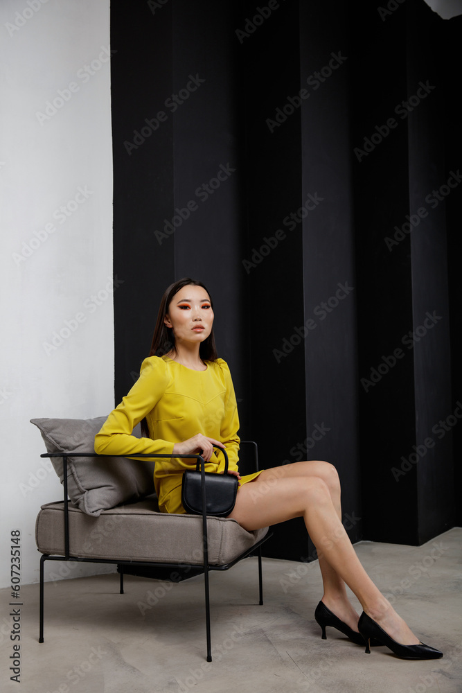 High fashion photo of beautiful elegant young asian woman in pretty yellow short dress, handbag, clutch posing on textured black and white wall. Slim figure. Model sits on chair
