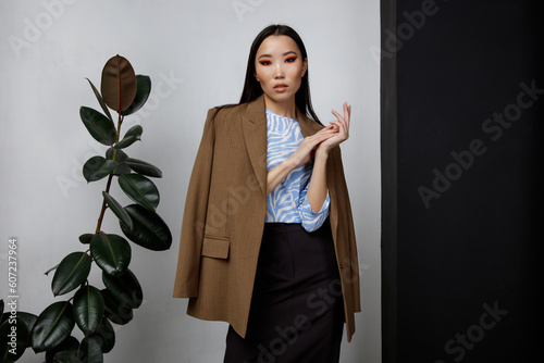 High fashion photo of beautiful elegant young asian woman in pretty brown beige jacket, blazer, blue top, long skirt posing on textured black and white wall, tall flower. Slim figure