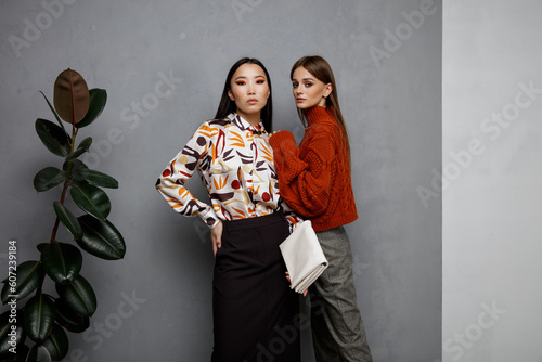 Two fashion models in orange beige outfits. Sweater, shirt, black skirt, gray pants, trousers, clutch, handbag. Beautiful young women. Asian and european girls posing on textured wall, tall flower
