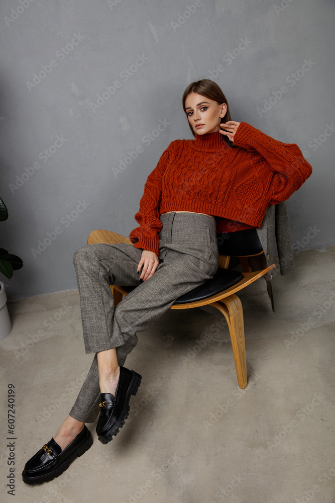 High fashion photo of beautiful elegant young woman in pretty orange terracotta sweater, gray pants, trousers posing on textured wall, tall flower. Slim figure. Model is sitting on a chair.