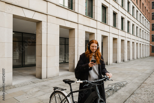 Cheerful redhead woman using mobile phone while standing with bicycle outdoors