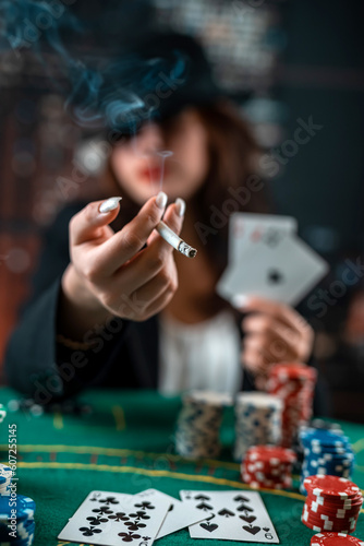 young girl stylishly dressed smokes a cigarette, blows smoke and plays cards on a table on a green cloth