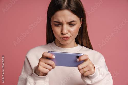 Young woman frowning while playing game on smartphone isolated over pink background