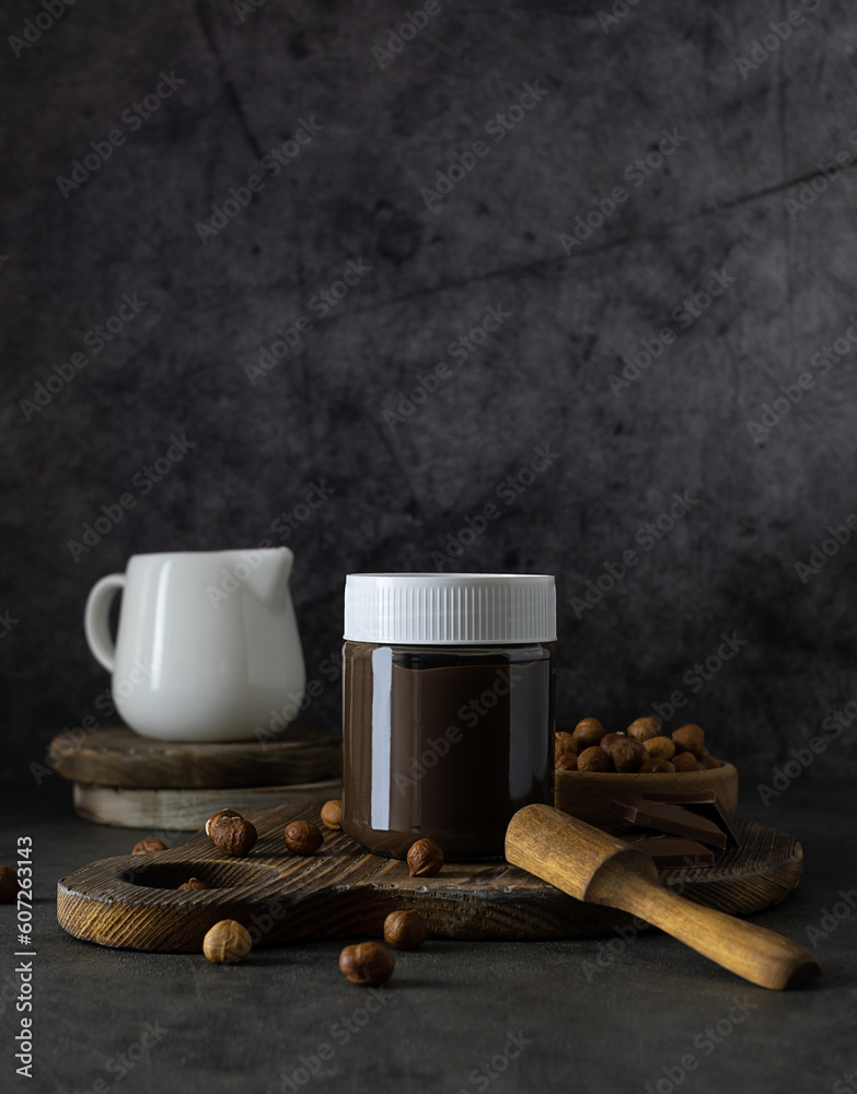 Nut and chocolate paste in a transparent jar with a white lid. A wooden spoon on the table. Nuts in a wooden bowl and white milkman in the background. Gray background