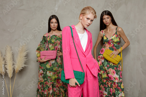 Three fashion models. Asians in identical looks, green dress with floral pattern, handbag, clutch. Blonde in pink fuchia suit, jacket, blazer, shorts. Beautiful young women. Sandy beige textured wall