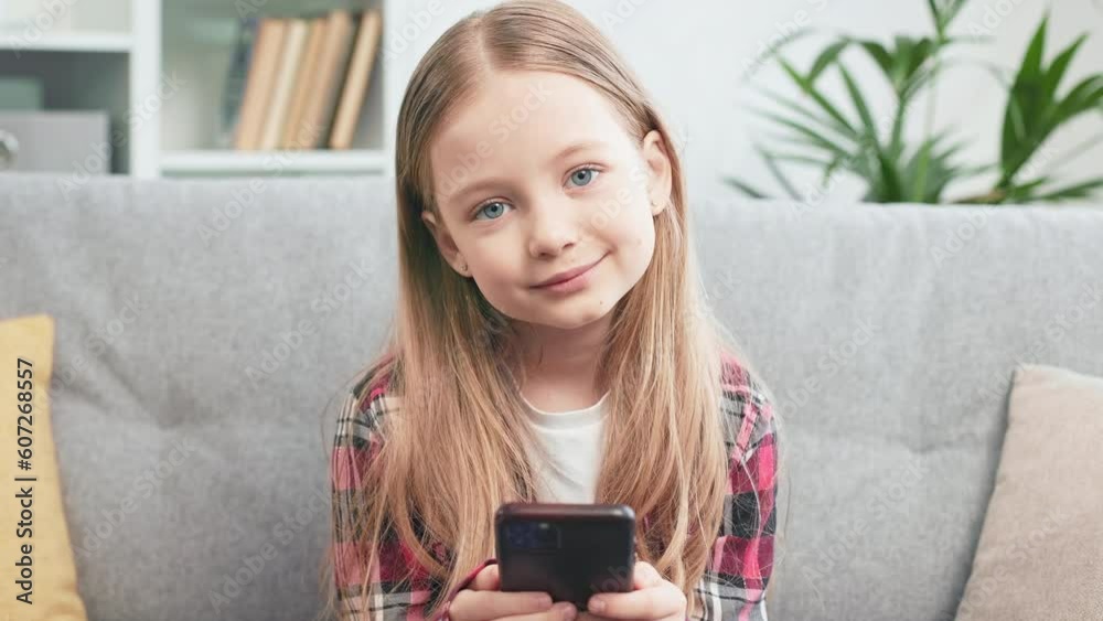 Charming little girl with wireless gadget in hands looking and smiling at camera. Positive blue-eyed child watching funny videos on personal smartphone and spending time at home.