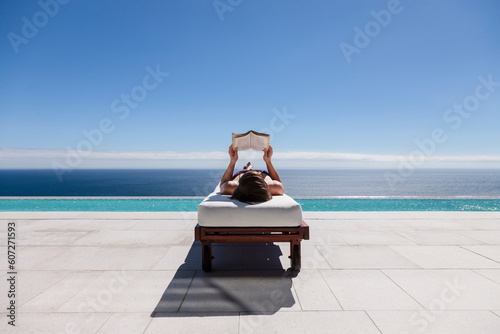 Billede på lærred Woman reading on lounge chair at poolside overlooking ocean