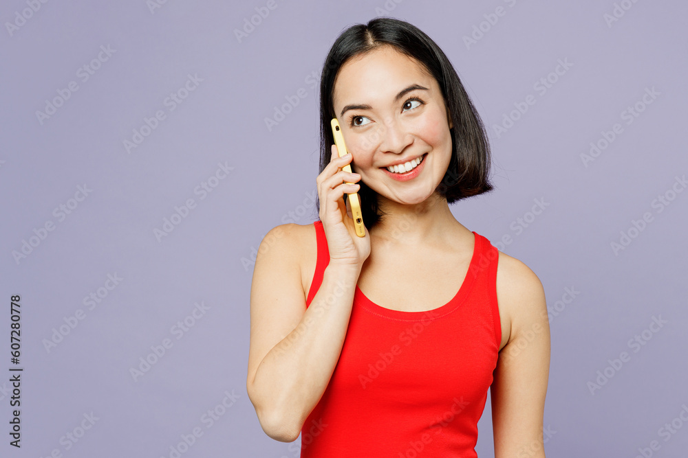 Young woman of Asian ethnicity she wear casual clothes red tank shirt talk speak on mobile cell phone conduct pleasant conversation isolated on plain pastel light purple background. Lifestyle concept. Young woman of Asian ethnicity she wear casual clothes red tank shirt talk speak on mobile cell phone conduct pleasant conversation isolated on plain pastel light purple background. Lifestyle concept.