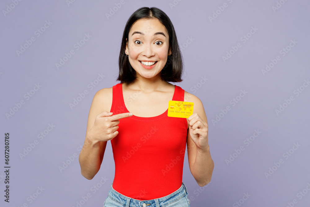 Young happy woman of Asian ethnicity she wear casual clothes red tank shirt hold point finger on mock up of credit bank card isolated on plain pastel light purple background studio. Lifestyle concept. Young happy woman of Asian ethnicity she wear casual clothes red tank shirt hold point finger on mock up of credit bank card isolated on plain pastel light purple background studio. Lifestyle concept.